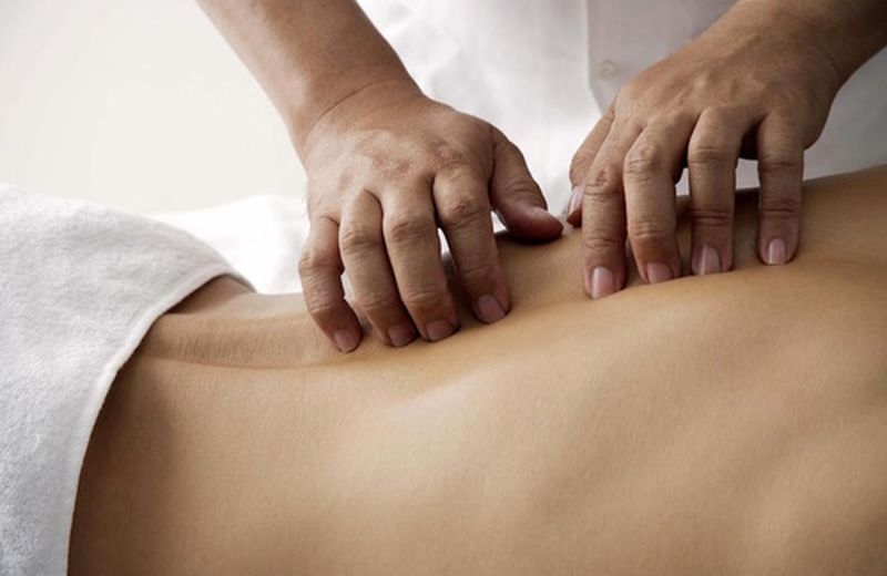 A woman is practicing acupressure by applying pressure with her fingertips on another person's back. The back is partially covered by a white towel, and the scene conveys a sense of relaxation and healing through therapeutic touch.