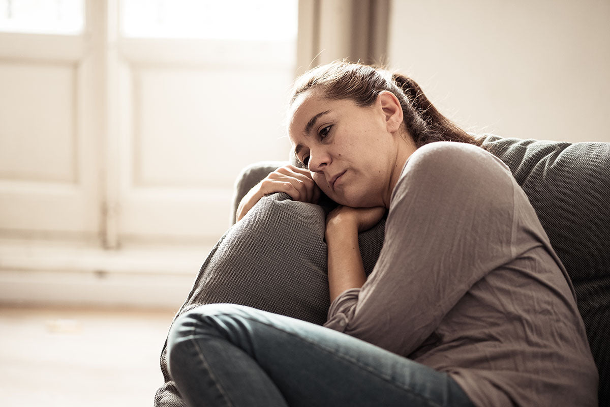 A woman appears pensive and worried while sitting on a couch, hugging her knees toward her chest. The expression on her face and the huddled posture suggests feelings of pain, anxiety, or depression, conditions often associated with fibromyalgia.