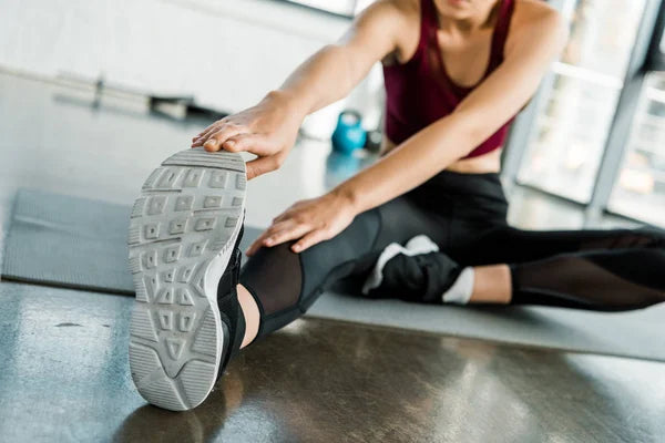 Woman in sportswear stretches by touching the toe of her sneaker, sitting on a mat in a gymnasium.