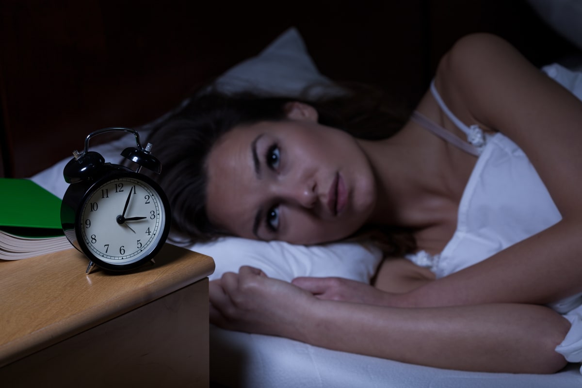 Woman lying in bed with restless expression next to an alarm clock, symbolizing insomnia.