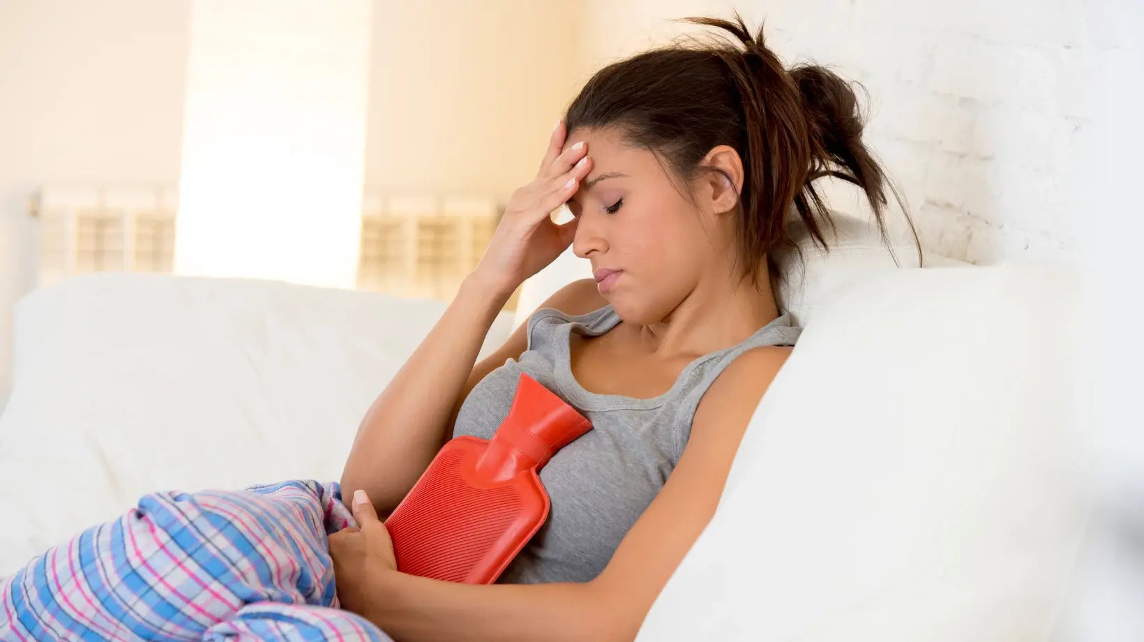 A woman sitting on the couch holds a hot water bottle on her belly with one hand and rests her forehead with the other, expressing discomfort that could indicate menstrual pain.