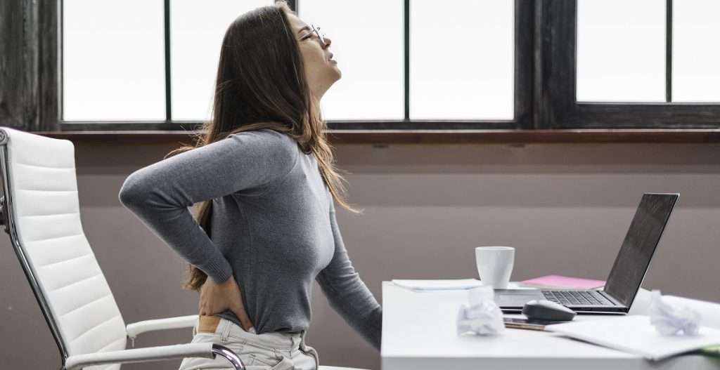 Woman in office with expression of pain while holding lower back, indicating possible back pain.