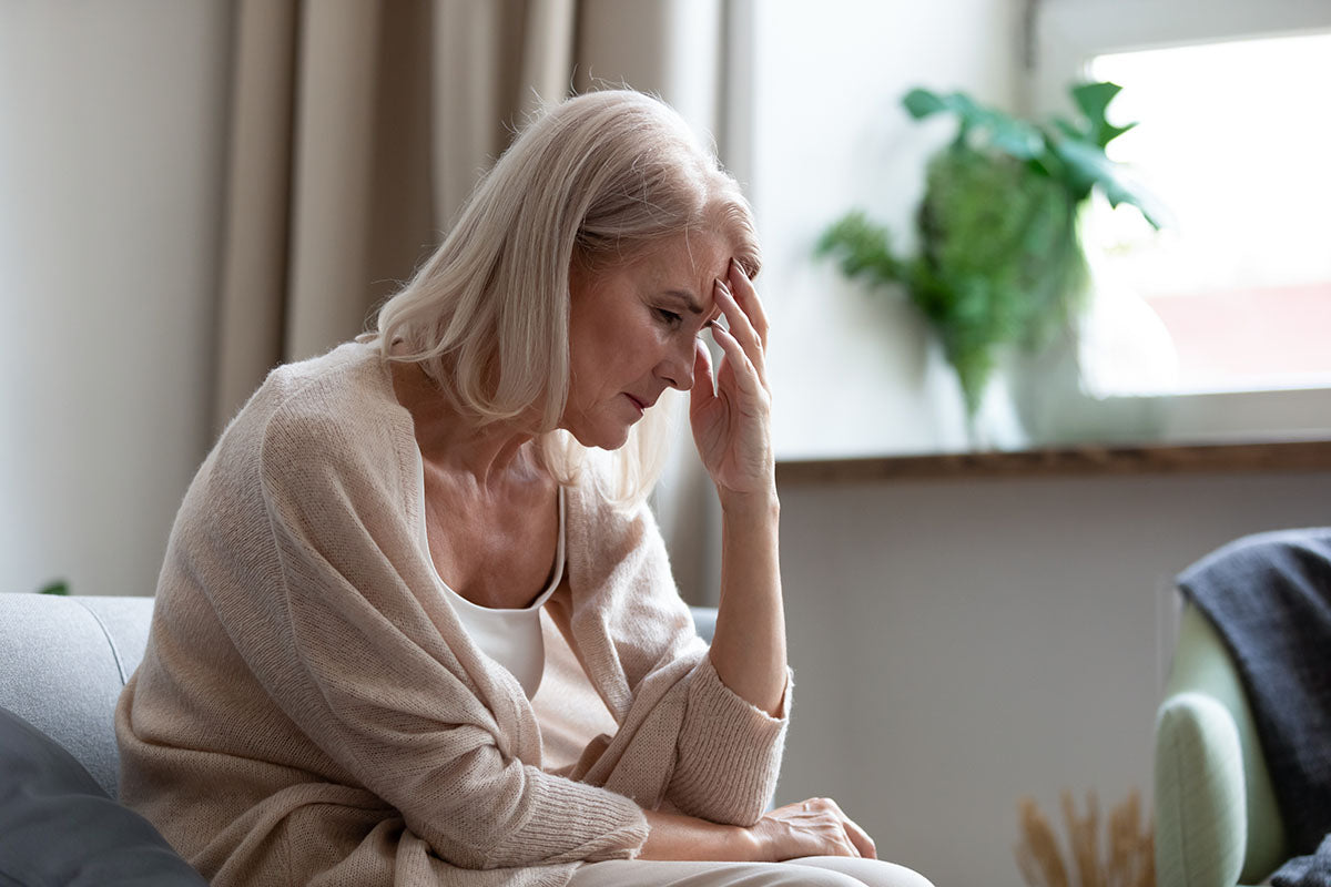 A woman with blond hair and beige robe sitting on a sofa, appearing apparently stressed and depressed, her left hand supporting her head in a gesture of despair or worry.