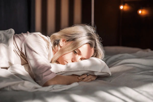 A woman appears thoughtful and absorbed as she lies on a bed with white sheets, resting her head on one arm and looking into the distance. The light filtering through the window illuminates her face, contributing to a depressed atmosphere.