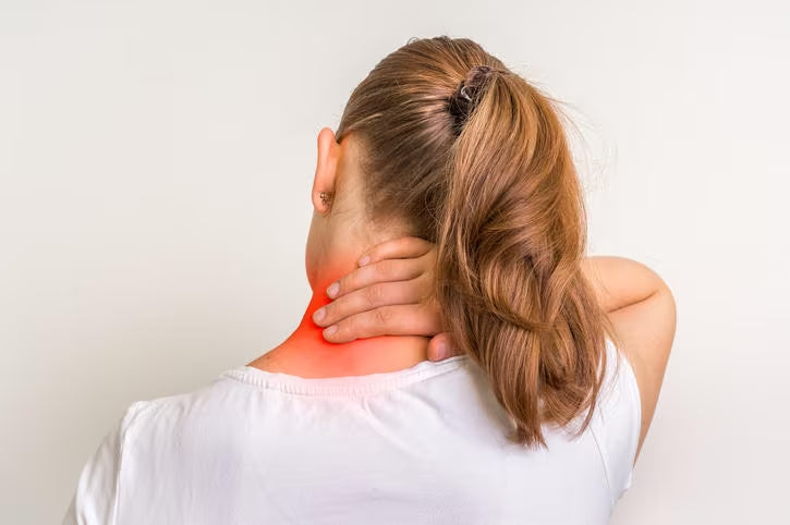 Image of a woman seen from behind holding the back of her neck with her right hand. There is an area highlighted in red on the neck that suggests pain or discomfort in that area.